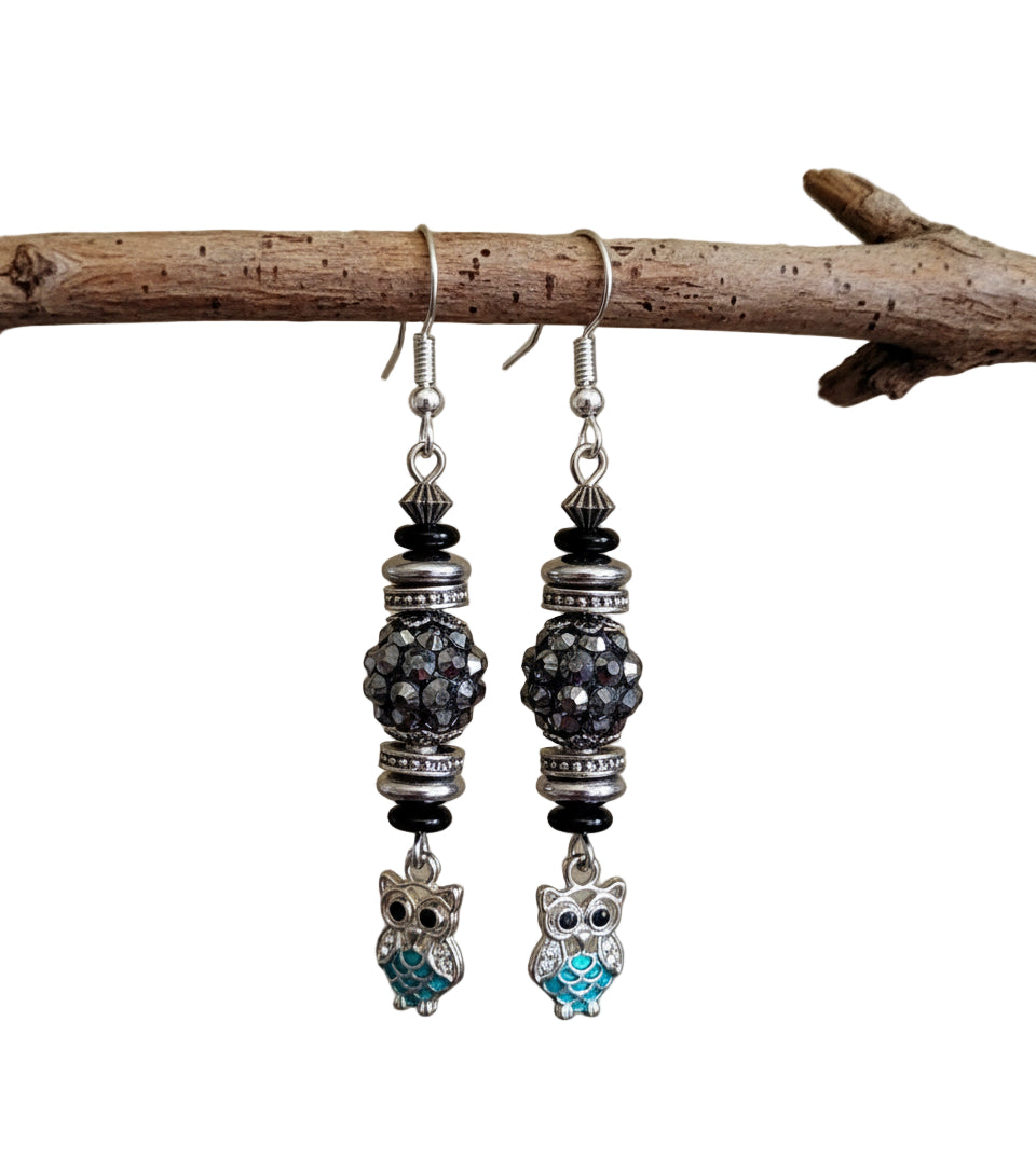 Pair of earrings with owl charms hanging from a branch on a white background