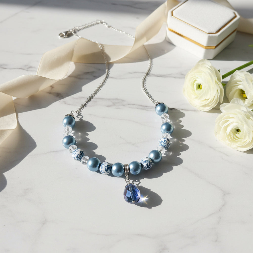 Blue beaded necklace with a teardrop pendant on a marble surface with flowers and a box.