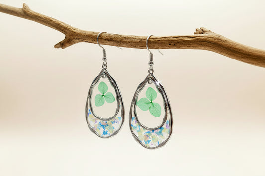Teardrop-shaped earrings with floral designs hanging on a branch against a beige background