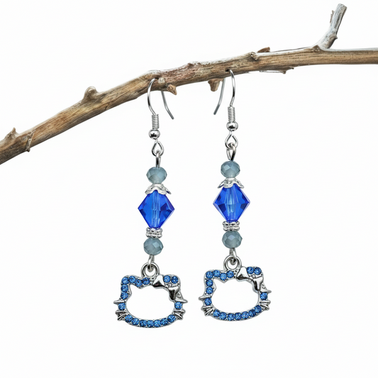 A pair of silver-plated cat charm earrings with blue gemstone or crystal beads, displayed against a marble background with blue and purple flowers.